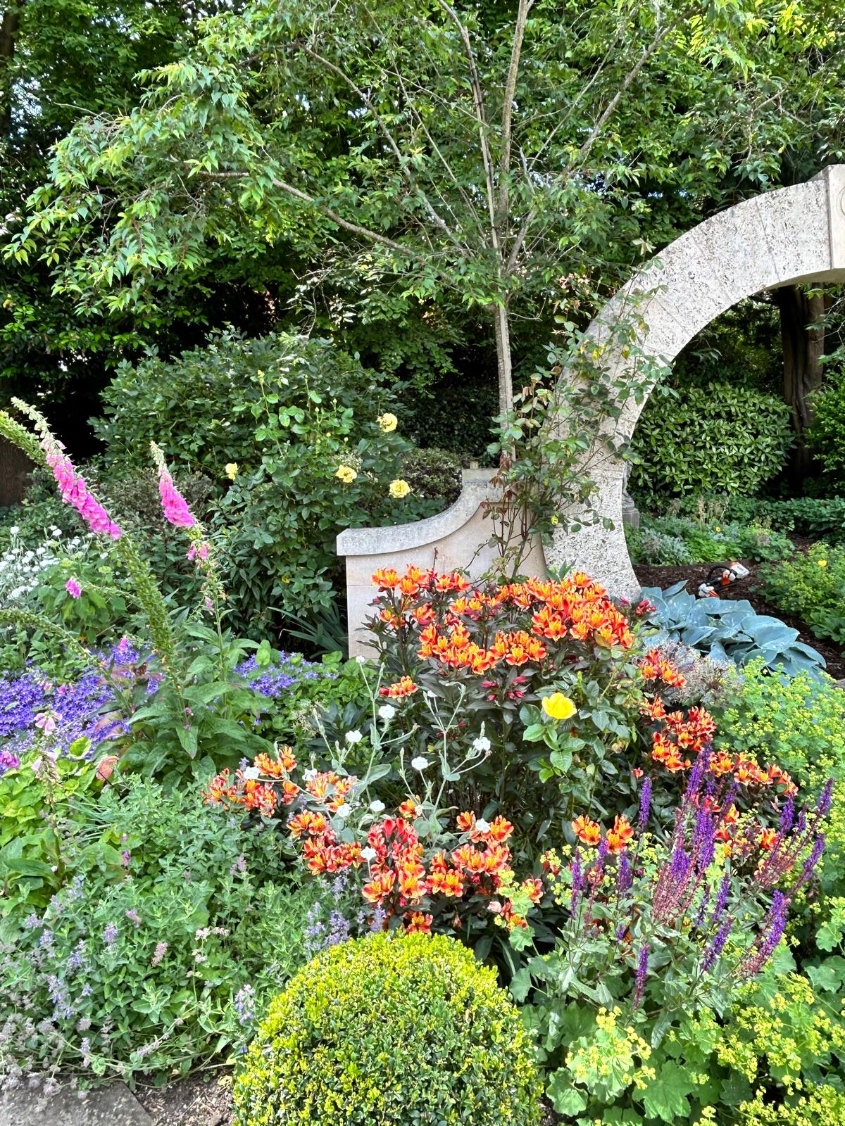 Layered flowering borders and leafy trees around a curved stone garden structure